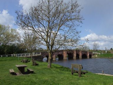 A riverside picnic area with wooden benches and tables near a stone bridge over a river at Island Yurt in Beckford near Tewkesbury