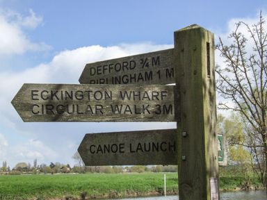 A wooden signpost with directions to Defford Bibingham Eckington Wharf and canoe launch in a grassy riverside area