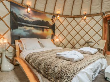 A bedroom with a large bed covered with a fur blanket and two robes on top a small table with two chairs and a landscape artwork on the wall at Island Yurt in Beckford near Tewkesbury
