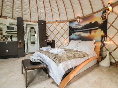 A bedroom with a double bed covered in white linens and a fur throw a small kitchenette area and a glass side table with a lamp at Island Yurt Beckford near Tewkesbury