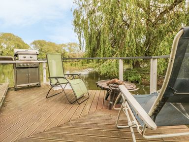 A wooden deck with two chairs a barbecue grill and a fire pit by a river at Island Yurt in Beckford near Tewkesbury