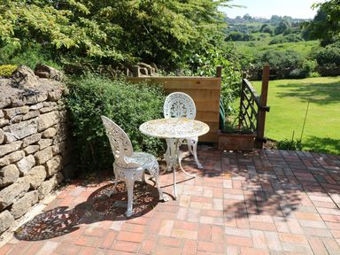 A patio with two white metal chairs and a round table near a stone wall and green bushes at The Loft at Lucott House in Bath
