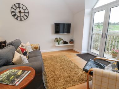A living room with a gray sofa with cushions a wall clock a wall-mounted TV and glass doors leading outside at The Loft at Lucott House in Bath