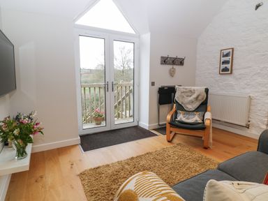 A living room with glass doors leading outside a wooden deck a chair with a cushion and throw and a TV mounted on the wall at The Loft at Lucott House in Bath