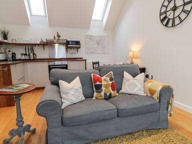 A living room with a grey sofa four pillows a side table and an open kitchen at The Loft at Lucott House in Bath