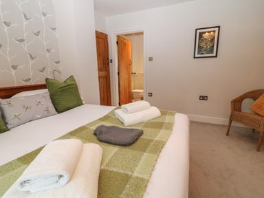 A bedroom with a bed covered by a green checkered blanket and towels on the bed a wicker chair in the corner and an open door revealing a bathroom at The Loft at Lucott House Bath
