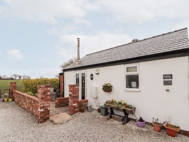 An exterior of a white cottage with a gray tiled roof and small brick patio with outdoor chairs surrounded by flower pots at The Old Stable in Mold