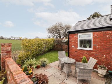 A small patio with a round glass table and two wicker chairs next to a red brick house and a grassy yard with plants at The Old Stable in Mold