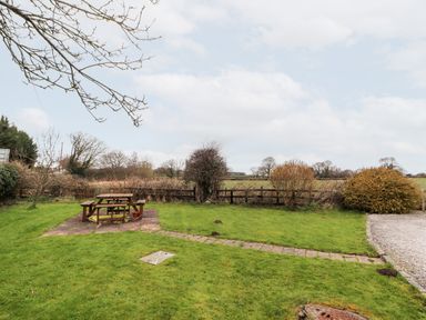 A grassy backyard with a wooden picnic table on a stone patio and some bushes near a wooden fence at The Old Stable in Mold