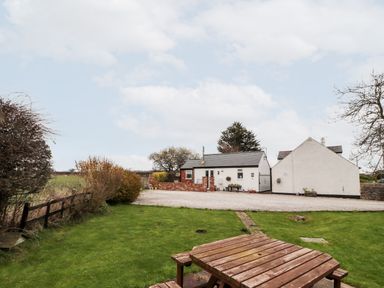 A garden with a wooden picnic table and benches and a gravel driveway leading to white buildings at The Old Stable in Mold