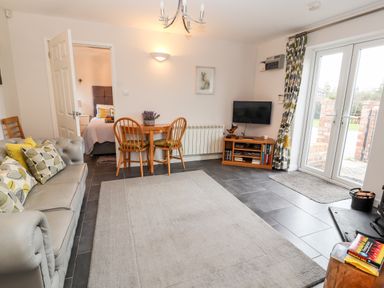 A living room with a gray sofa and patterned cushions a wooden table with two chairs a small wooden TV stand with a television and glass doors leading outside at The Old Stable in Mold