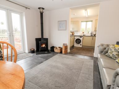 A living room with a wood stove near a door and a kitchen with a washing machine seen through an opening at The Old Stable in Mold