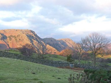 A grassy field with stone walls and leafless trees with mountains in the background at High View Lodge White Cross Bay near Troutbeck Bridge