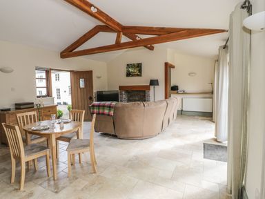A dining area with a wooden table and four chairs next to a living room with a curved beige sofa a fireplace and wooden ceiling beams at The Old Dairy in Newnham-on-Severn