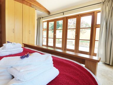 A bedroom with a wooden bed frame folded towels on a red blanket wooden wardrobes and glass doors with curtains at The Old Dairy in Newnham-on-Severn
