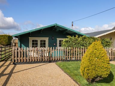 Exterior of a green wooden cabin with a fenced patio and nearby shrubs at The Nook in Royal Wootton Bassett