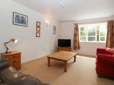 A living room with a wooden coffee table a red armchair a black sofa a wooden side table with a lamp and a TV on a wooden stand under a window at Wheelbarrow Cottage in Stoke Prior near Leominster