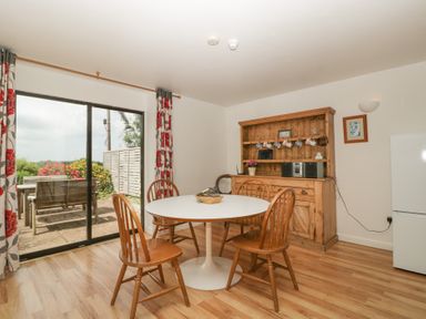 A dining area with a round white table surrounded by wooden chairs and a wooden cabinet with hanging mugs at Wheelbarrow Cottage in Stoke Prior near Leominster