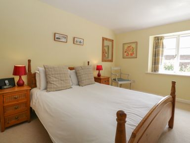 A bedroom with a wooden bed, two bedside tables with red lamps, a chair, window with curtains and wall art at Wheelbarrow Cottage in Stoke Prior near Leominster