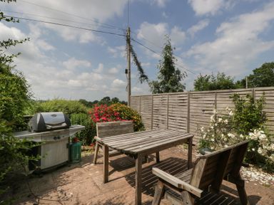 An outdoor patio with wooden table and chairs a barbecue grill and flowers at Wheelbarrow Cottage Stoke Prior near Leominster