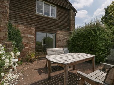 An outdoor patio with wooden table and chairs next to a brick and timber house with sliding glass door and plants at Wheelbarrow Cottage in Stoke Prior near Leominster