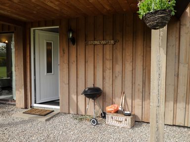 A wooden porch with a white door a small charcoal grill a wicker basket with firewood tools and a hanging basket of flowers at Stone Water Cottage Strathpeffer