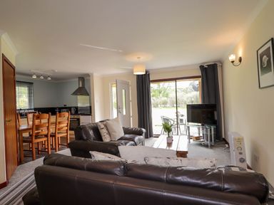 A living room with leather sofas a wooden dining table and chairs a kitchen area and a television near a sliding glass door at Stone Water Cottage in Strathpeffer