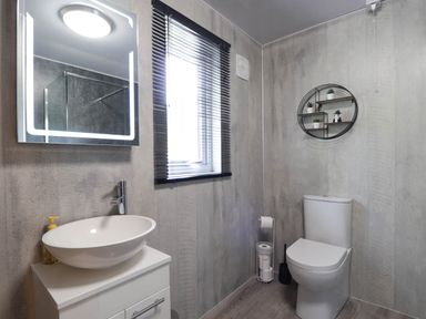 A bathroom with a toilet a sink a backlit mirror a window with blinds and a round wall shelf in Stone Water Cottage Strathpeffer