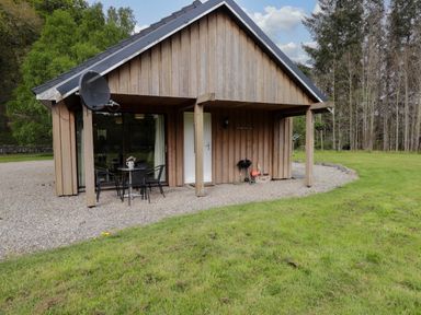 A wooden cabin with a small patio table and chairs on a gravel area surrounded by grass at Stone Water Cottage in Strathpeffer
