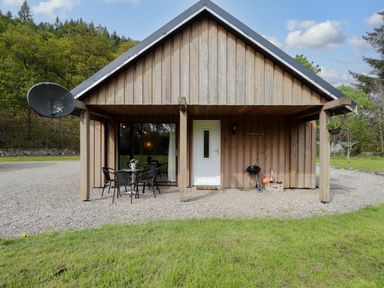 A wooden cabin with a white door and patio table with chairs on a gravel area at Stone Water Cottage in Strathpeffer