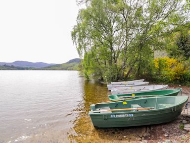 Several boats on the shore of a lake with trees and hills in the background at Stone Water Cottage in Strathpeffer