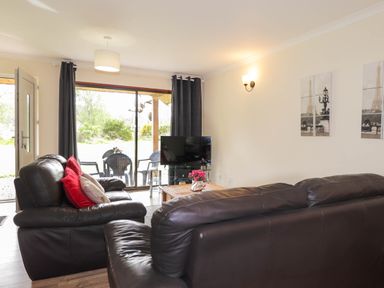 A living room with leather sofas a coffee table a television and a sliding glass door leading to an outdoor seating area at Pebbles Cottage in Achilty near Strathpeffer