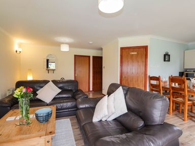A living room with leather sofas wooden coffee table flowers and dining area with wooden chairs at Pebbles Cottage in Achilty near Strathpeffer