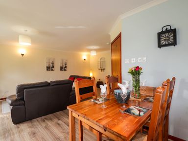 A dining area with a wooden table set for four and a living room with leather sofas in an open plan space at Pebbles Cottage in Achilty near Strathpeffer