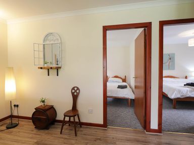 A hallway with a wooden chair small cabinet and floor lamp with two bedrooms visible through open doorways at Pebbles Cottage in Achilty near Strathpeffer