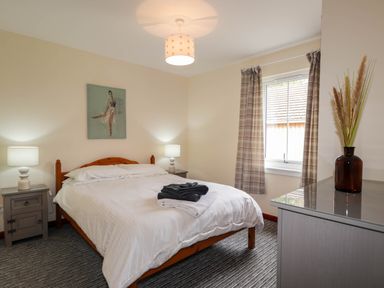 A bedroom with a bed with white bedding two bedside tables with lamps a window with plaid curtains a grey dresser and a painting above the bed at Pebbles Cottage in Achilty near Strathpeffer