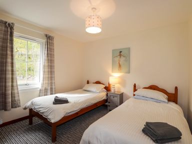A bedroom with two single beds a nightstand with a lamp and a window with plaid curtains at Pebbles Cottage in Achilty near Strathpeffer