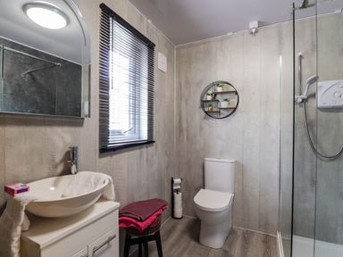 A bathroom with a toilet a sink with a mirror a stool with towels a window with blinds a shower and wall shelves with small plants at Pebbles Cottage Achilty near Strathpeffer