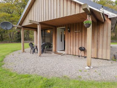 An outdoor area with wooden cabin wall a door a small table with chairs a hanging flower basket and a charcoal grill at Pebbles Cottage in Achilty near Strathpeffer