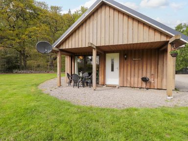 A wooden cottage with a white door patio table with chairs and a barbecue on a gravel area at Pebbles Cottage in Achilty near Strathpeffer