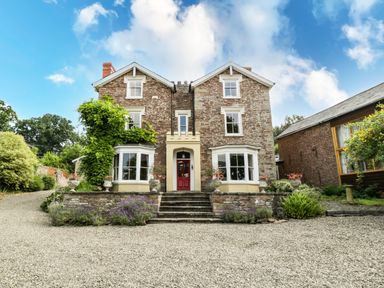 A two-story stone house with a red door and two bay windows surrounded by plants and gravel driveway at Wheelbarrow Castle in Leominster
