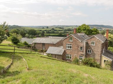 A stone house with multiple chimneys and white windows surrounded by green fields and trees at Wheelbarrow Castle in Leominster