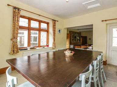 A dining room with a large wooden table surrounded by chairs near a window with patterned curtains at Wheelbarrow Castle in Leominster