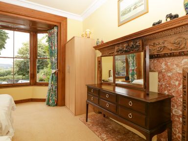 A bedroom with a wooden dresser with mirror beside a wardrobe and a large bay window at Wheelbarrow Castle in Leominster