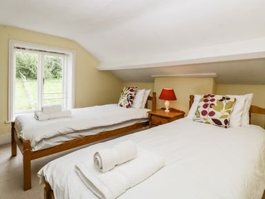 A bedroom with two single beds white linens a wooden nightstand with a red lamp and a window showing greenery at Wheelbarrow Castle in Leominster