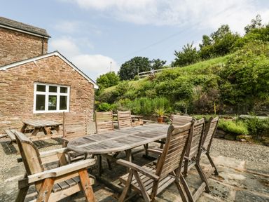 An outdoor patio with a wooden dining table and chairs near a stone house and garden at Wheelbarrow Castle in Leominster