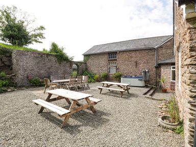 An outdoor patio area with wooden picnic tables a dining table with chairs stone walls and a hot tub at Wheelbarrow Castle in Leominster