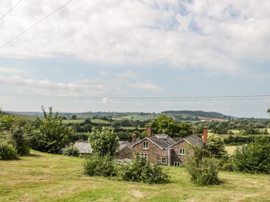A countryside house with brick walls surrounded by trees and bushes with fields and hills in the background at Wheelbarrow Castle in Leominster