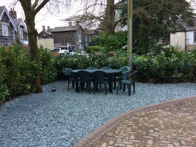 A garden area with plastic chairs and a table on gravel near trees and houses at Sandown in Bowness-on-Windermere