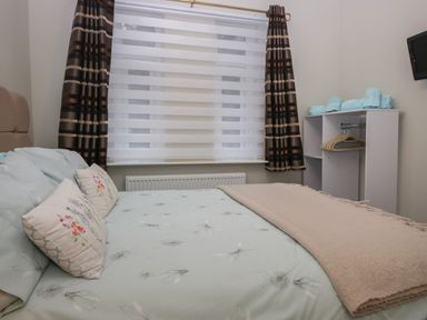 A bedroom with a bed a window with curtains a small shelving unit with towels and hangers and a wall-mounted TV at Sandown in Bowness-on-Windermere